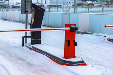 Outdoor barrier arm system on a snowy road with a barrier arm down, equipped with a red light, with various buildings in the background, Customer access control parking lot