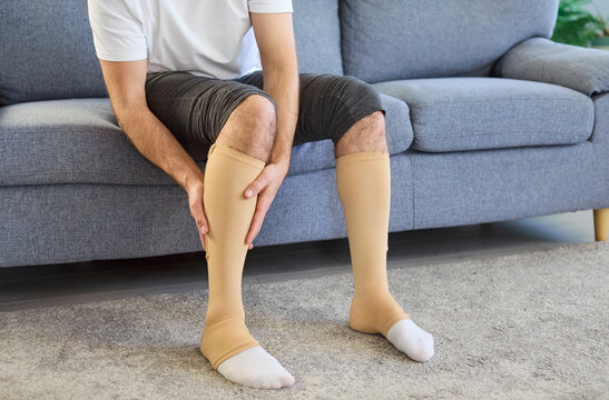 Man in medical compression socks massaging painful calves. Beige hosiery supports legs and varicose veins at home improving circulation and easing swelling. Thrombosis risk and venous therapy.