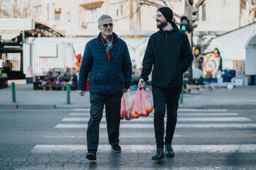 Fototapeta premium Two men walk across a busy urban crosswalk on a chilly day. One older in a blue jacket and scarf, the other younger in black, carrying pink bags from a market.