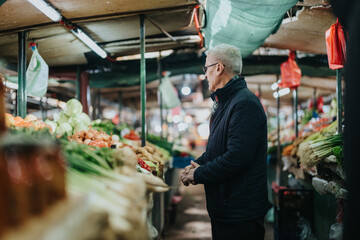 An elderly man in a dark jacket surveys fresh produce at a bustling indoor market, conveying...