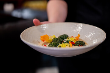 hand holding a white, rustic bowl filled with elegantly presented gourmet food. The dish features dark green ingredients, likely leafy greens or pasta, yellow and orange accents, and a creamy sauce