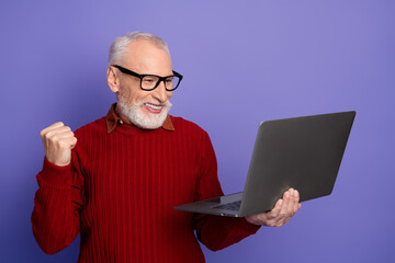Mature man with grey hair wearing red sweater smiles while holding laptop against purple background