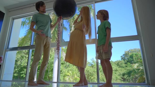 Happy family celebrating indoor gender reveal party at home. Pregnant mother pops balloon with blue confetti announcing it's a boy, while father and elder brother hug, cheer and enjoy joyful family - Powered by Adobe