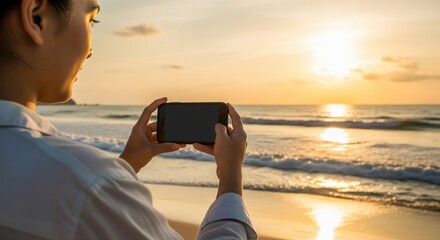 Woman holding a smartphone to capture the beautiful ocean sunset on a tropical beach.