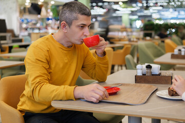 A man in a yellow sweater sips coffee from a red cup at a trendy cafe, surrounded by colorful decor, creating a cozy, vibrant atmosphere