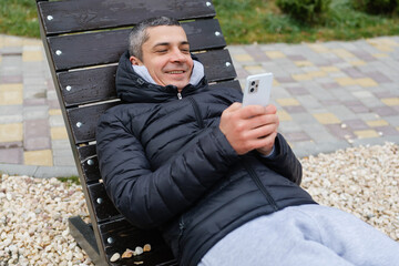 A man lies back on a lounge chair, enjoying a sunny day in the park. He checks his smartphone, smiling and relaxed among nature