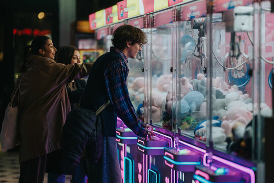 Three teens lean toward the prize claw, smiling as they guide the lever in a neon-bright arcade, chasing soft plush toys and sharing a playful, energetic moment.