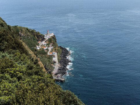 Lighthouse Farol do Arnel seen from a viewpoint, with rocky coastline, winding road and coastal houses along the Azorean shore.