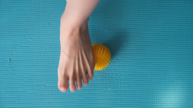 Female feet are doing foot exercises by rolling a prickly massage yellow ball under the foot on the yoga mat pressing down in different sides. Training to correct and relax the foot fascia. 4k footage