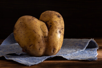 A uniquely shaped heart-forming ugly potato rests on a blue cloth against a dark wooden background.