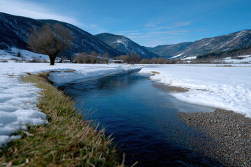 A pristine river gently flows through a tranquil snowy landscape, illuminated by soft winter sunlight, highlighting the serene beauty of nature during this calm season.