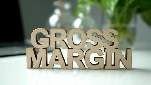 Close-up on wooden 'Gross Margin' letters placed on a white table, set against a blurred background featuring a plant and a laptop.