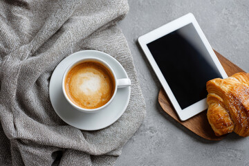 Overhead shot of a coffee cup, baked treat, and tablet with white screen on a textured concrete surface