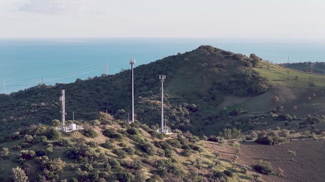 Aerial view of cell towers stand tall on a lush green hill, contrasting against the clear blue sea in the background, Rocca Imperiale, Calabria, Italy.