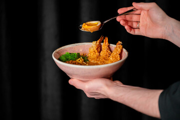 A hand adds a soft-boiled egg to a bowl of tempura shrimp ramen, ready to be eaten