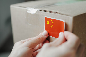 Close-up of hands placing a sticker of the Chinese flag on a cardboard shipping box. Concept of international delivery, export and import from China, logistics, e-commerce and global trade.