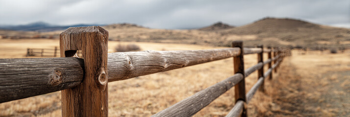 A weathered wooden fence lines the path to a rustic barn on a ranch, surrounded by open fields and distant mountains under gray skies, capturing the peaceful rural atmosphere, banner
