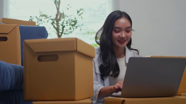 Smiling Asian woman typing on a laptop computer while sitting on the floor among cardboard boxes. Relocating to a new home