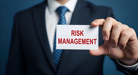 Obraz premium Businessman in dark suit holds small white card displaying bold red text reading risk management against a deep blue background