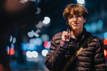 A young man wearing a quilted jacket and glasses stands outdoors on a city street at night, with bright bokeh lights in the background, conveying a calm urban mood.