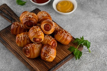 Mini hot dog bites with mustard and ketchup. Sausages baked in a yeast dough cover. Pigs in a blanket on a wooden board on a gray background. Top view, copy space.