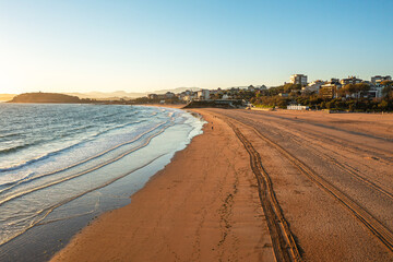 Drone view of wide sandy beach with gentle waves, morning sunlight, seaside and distant city skyline