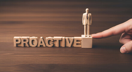 Wooden blocks spell proactive with a small businessman figure standing atop the final block as a finger gently pushes the base on a dark wood surface