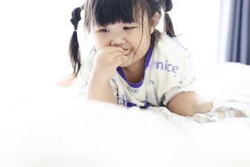Fototapeta premium A cute little Asian girl is lying in bed eating cookies,Sitting on the bed, eating cookies and drinking milk. Cute little girl in casual clothes is indoors at home at daytime.
