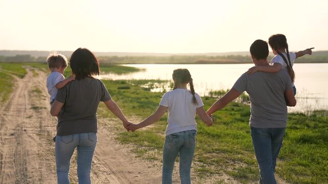 Family walking hand in hand along dirt road by lake at sunset with parents carrying child and sibling holding hand near green meadow and dog nearby parent mother father child daughter son walk lake