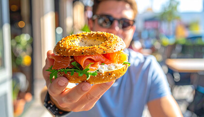A person holding a delicious bagel sandwich filled with smoked salmon, cream cheese, and fresh arugula, presented towards the camera in an outdoor setting.
