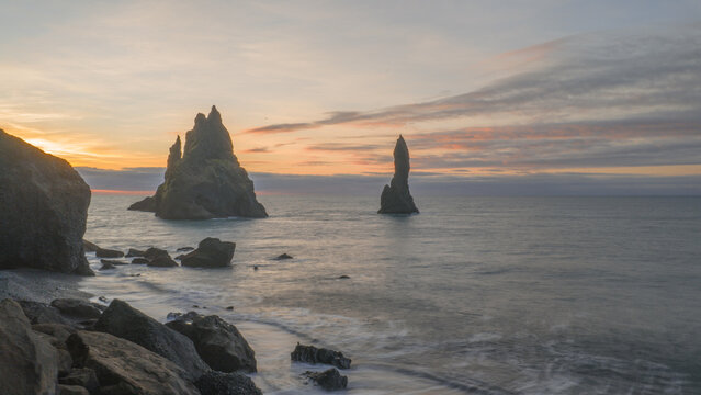 Aerial view of jagged basalt sea stacks rise dramatically from the churning Atlantic waters against a canvas of fiery sunset hues, Reynisfjara, Myrdalshreppur, Iceland.