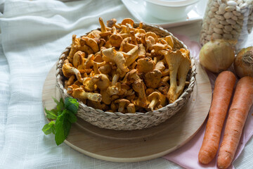 A full basket of chanterelles displayed on a wooden board with carrots, onions, and mint. Soft daylight enhances textures, creating a warm, rustic atmosphere suited for cooking, seasonal harvest, and