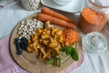 A rustic food arrangement featuring white beans, carrots, chanterelles, blueberries, and green herbs placed on a wooden board with glass jars of lentils and grains in the background. Perfect for recip