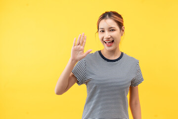 Friendly young asian woman saying hi and waving with a cheerful smile on yellow background.