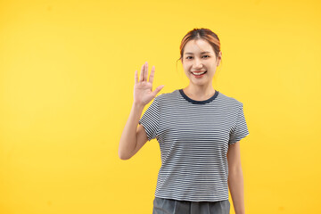 Friendly young asian woman saying hi and waving with a cheerful smile on yellow background.