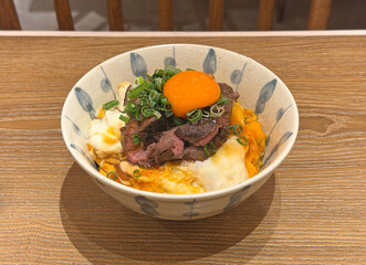 A close-up of a savory beef bowl topped with a glistening raw egg yolk and fresh green onions, presented in a decorative bowl on a textured wooden surface