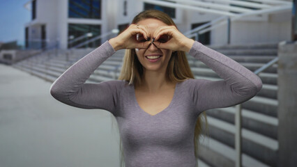 Woman outdoors mimicking binoculars with hands on stairs background smiling building.