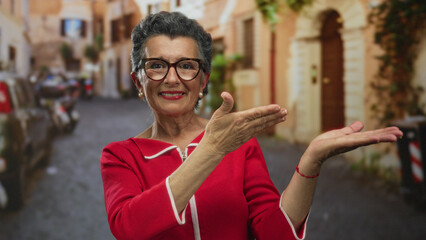 Senior woman with grey hair extends hands in an old town street, wearing red, smiling confidently...