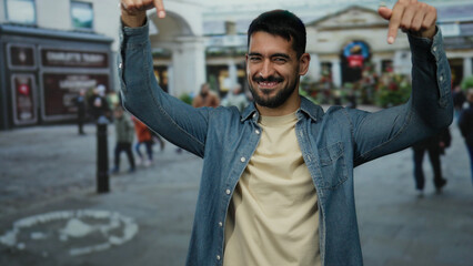 Young man pointing down with a smile in an outdoor urban street setting with blurred buildings and...