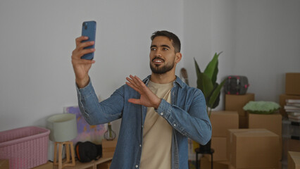 Young man making a video call in a new home surrounded by moving boxes, wearing a casual shirt, expressing happiness with open arms in a cozy living room.