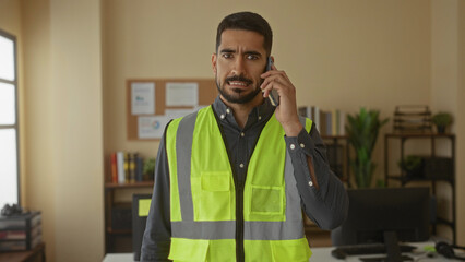 Young man wearing safety vest talks on phone in a modern office setting, highlighting workplace communication and professional attire.