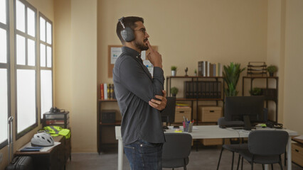Young man wearing headphones in an office setting, surrounded by desks, computers, and potted plants, engaged in conversation, suggesting a modern workplace environment.