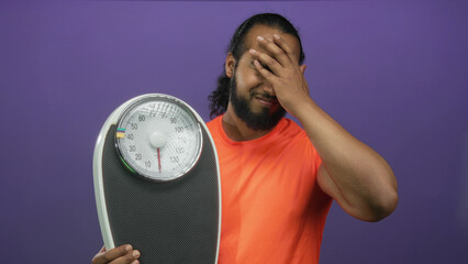 Man holding a bathroom scale and pressing hand to forehead in purple studio; frustration weight concern.