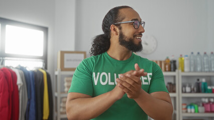 Man in volunteer shirt claps hands in a donations building while smiling and organizing supplies on shelves; community support gratitude.