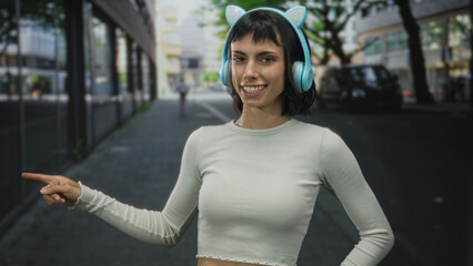 Woman with turquoise cat ear headphones points finger to the left along a tree lined street in daylight; playfulness.
