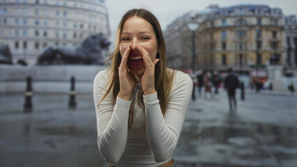 Young woman cupping hands to mouth shouting in a street plaza by lion sculptures and historic building, wet pavement and passing pedestrians; joy.
