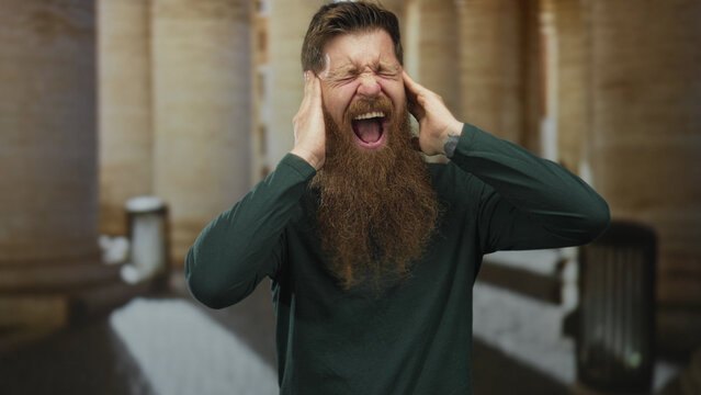 Man with long beard screaming with hands raised by historic stone building entrance; anger release solitude.