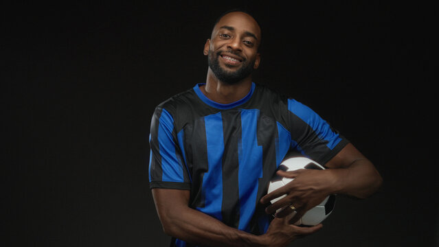 Man soccer player smiling holding soccer ball with bare forearm visible wearing blue and black jersey in black studio; team pride. - Powered by Adobe