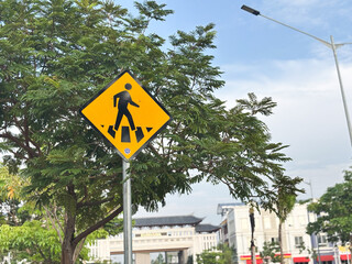 A bright yellow, diamond-shaped warning sign depicting a pedestrian crossing is mounted on a metal pole, partially framed by the lush green foliage of a tree on a sunny day.
