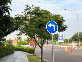 A blue, circular mandatory road sign indicating a compulsory left turn is prominently displayed on a metal pole next to a paved walkway and lush green landscaping.